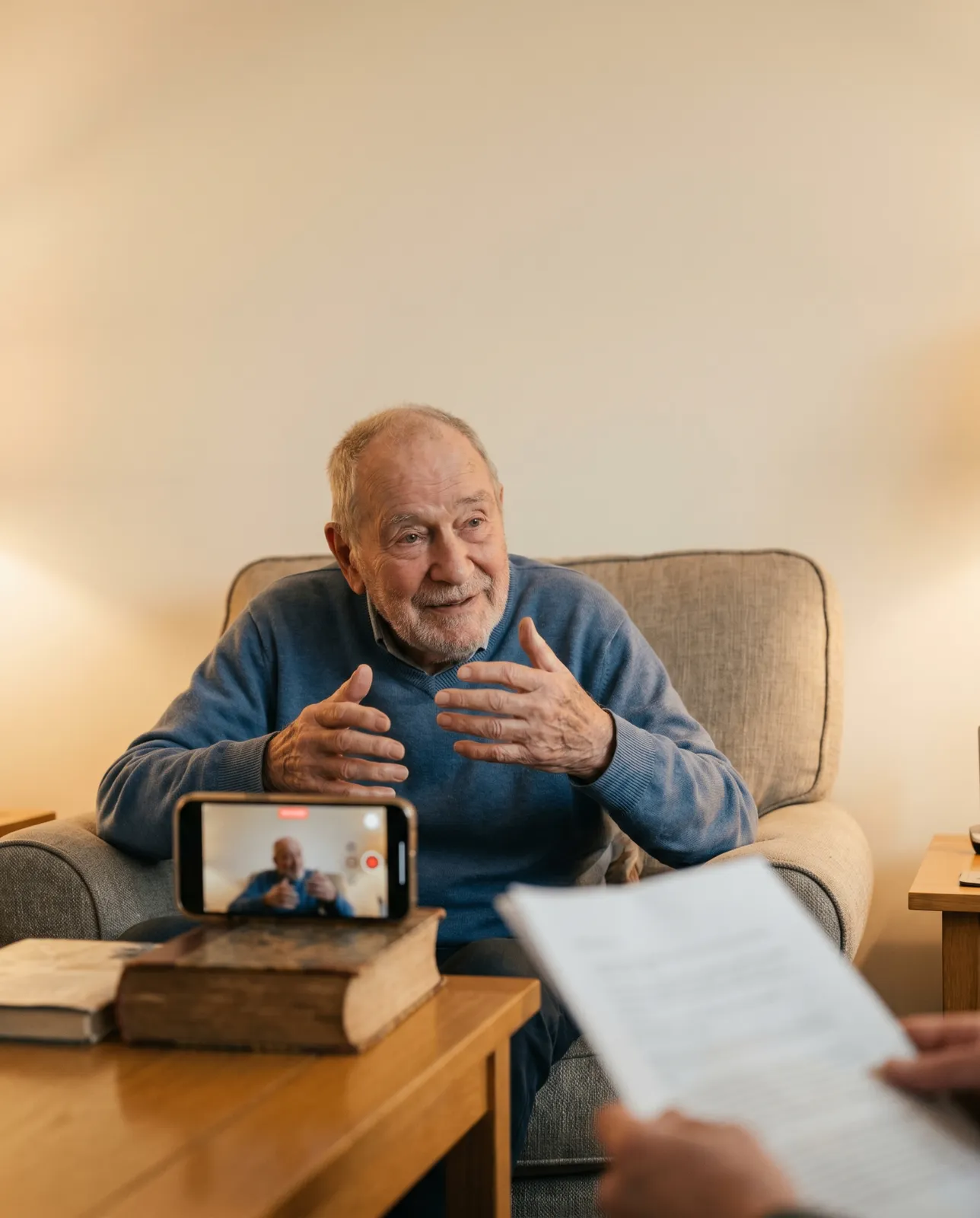 An older man in a blue jumper sitting in his armchair, hands open mid-story, with a phone propped on a stack of old books recording him — a grandchild's question sheet visible in the foreground.